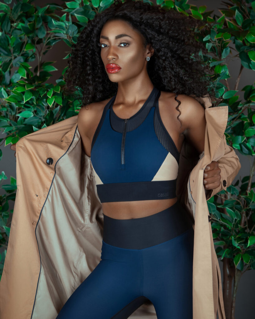 Black woman in blue fitness outfit with brown trench coat posing by green leaf background.