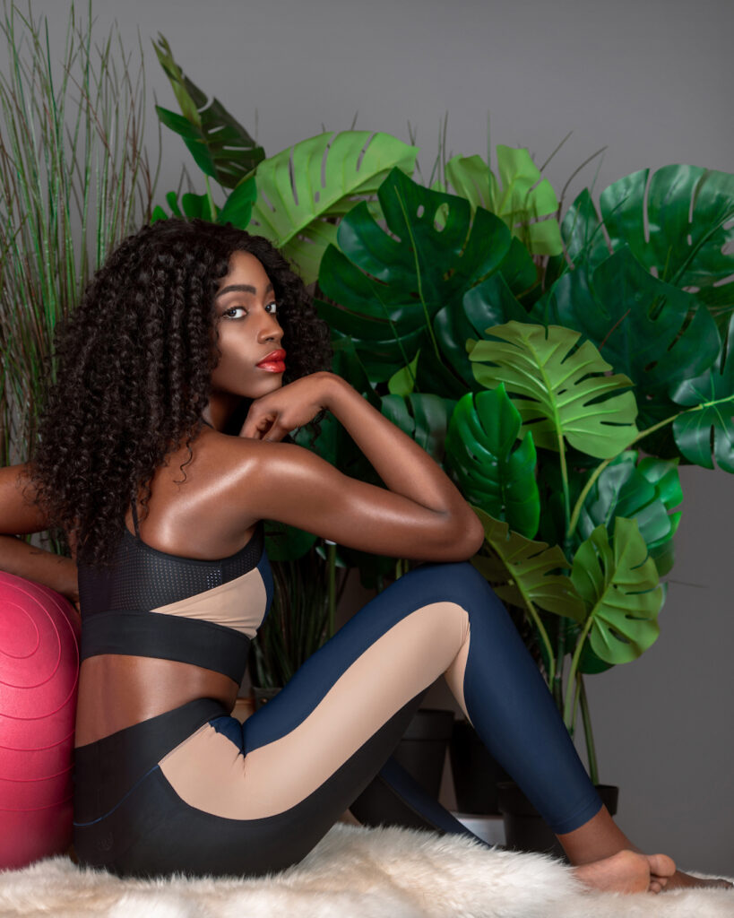 Black woman in blue fitness outfit leaning on a physio ball in grey studio.