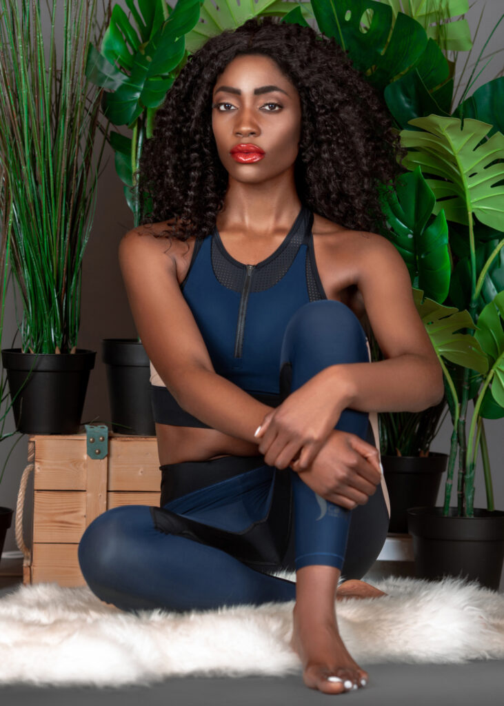 Portrait of a beautiful young black woman with long curly black hair, beautiful makeup sitting by herself on fur in a studio with a plant background wearing a blue fitness outfit looking at the camera.