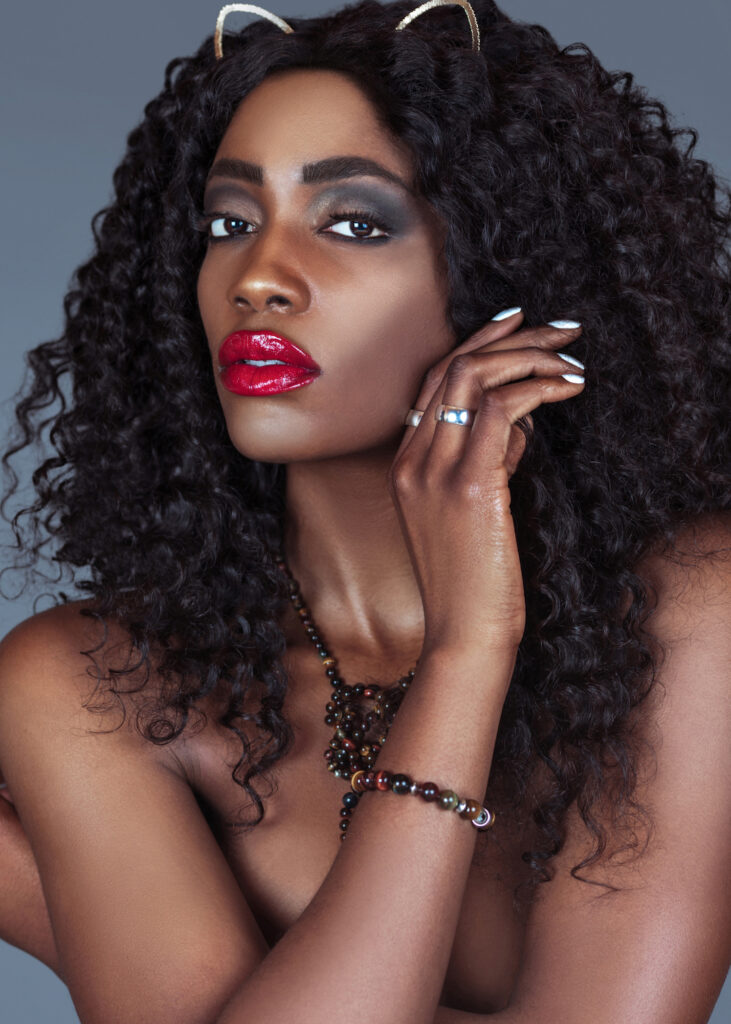Portrait of a sensual black cat woman with long curly black hair, beautiful red lips and eye shadow posing by herself in a studio with a grey background just wearing jewelry and a necklace.