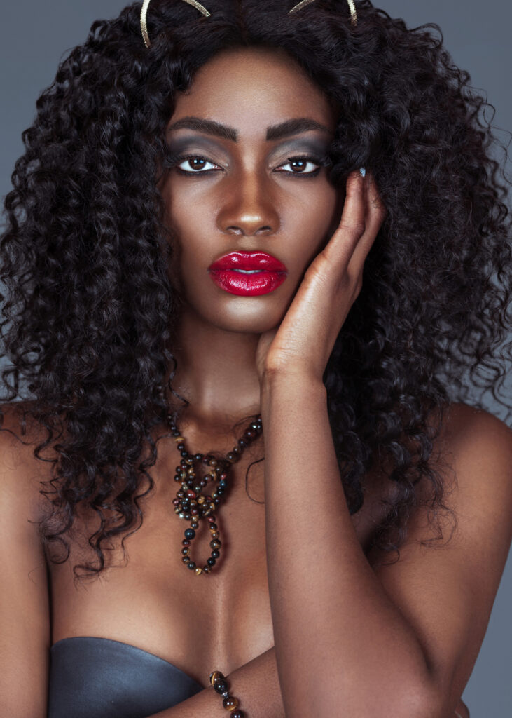 Spring Is In The Air. Black woman with curly hair, red lips and eyeshadow wearing only jewelry and necklace in studio with grey background.