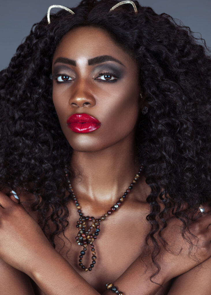 Portrait of an elegant black cat woman with long curly black hair, beautiful red lips and eye shadow posing by herself in a studio with a grey background just wearing jewelry and a necklace.