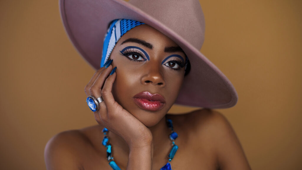 Sensual young black female with gorgeous makeup posing by herself inside a studio wearing a grey hat, blue head scarf & jewelry.