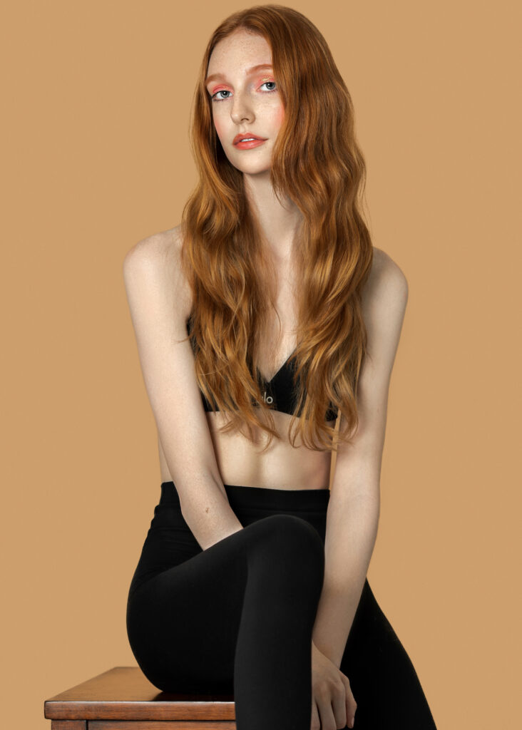 Modern beauty and self-expression. Brisbane with long red hair, natural makeup, sitting on a table wearing black top and leggings, studio with pecan background.