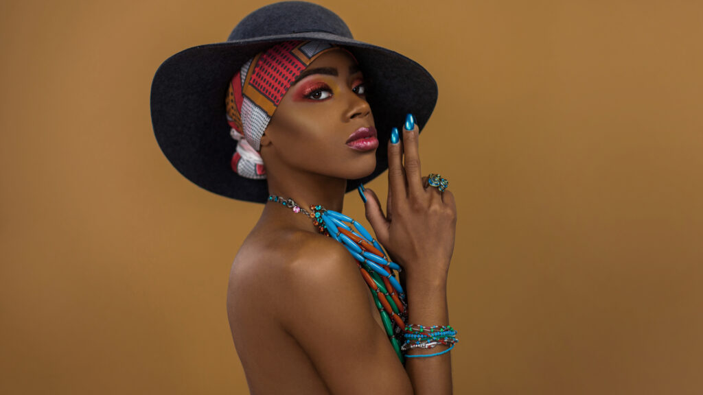 Fierce young black female with gorgeous makeup posing by herself inside a studio wearing a grey hat, blue head scarf & jewelry.