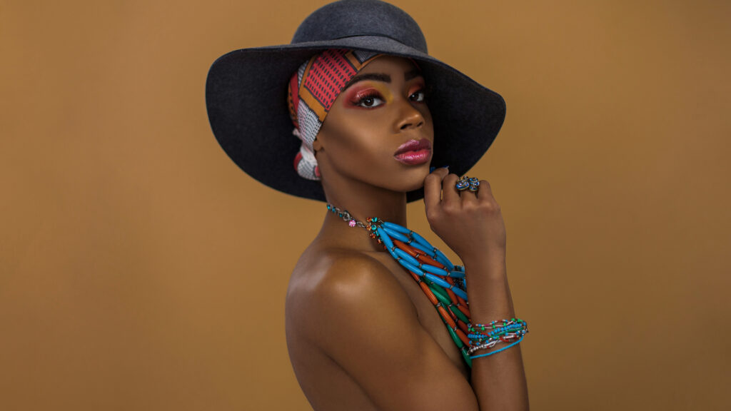 Elegant young black female with gorgeous makeup posing by herself inside a studio wearing a grey hat, blue head scarf & jewelry.