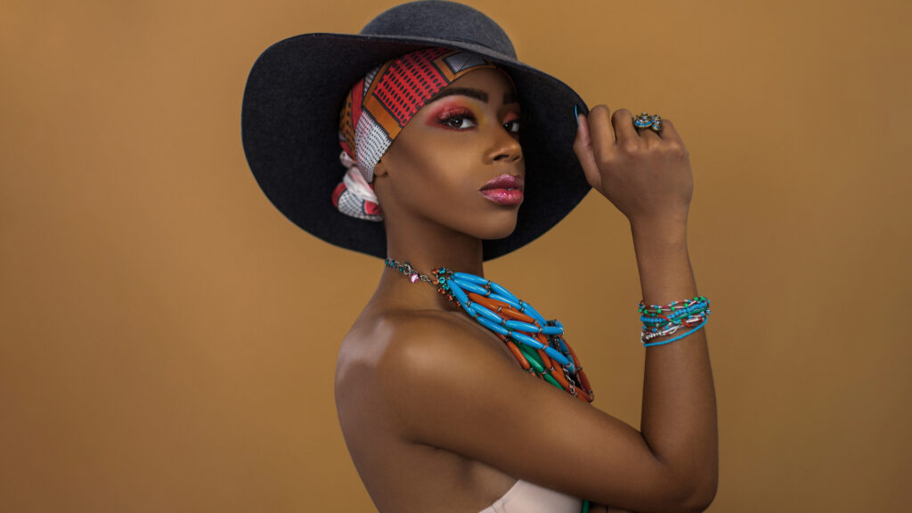 Sophisticated young black female with gorgeous makeup posing by herself inside a studio wearing a grey hat, blue head scarf & jewelry.
