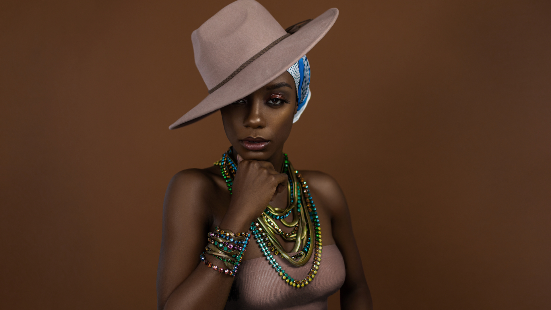 Gypsy Queens Editorial. Fierce looking young black female with beautiful makeup posing by herself in a studio wearing a beige strapless top, head scarf, beige hat and gorgeous colorful jewelry.