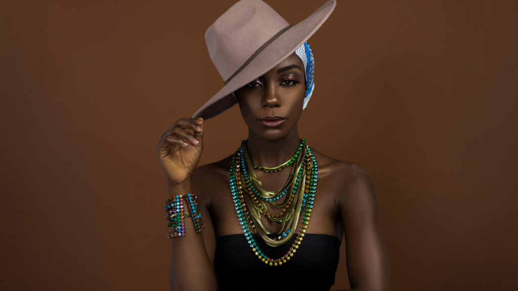 A fierce looking young black female with beautiful makeup posing by herself in a studio wearing a black strapless top, white and blue head scarf, beige hat and gorgeous colorful jewelry.