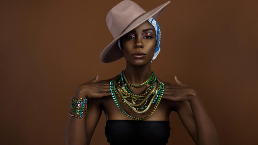 Sensual looking young black female with beautiful makeup posing by herself in a studio wearing a black strapless top, blue & white head scarf, brown hat and gorgeous colorful jewelry.