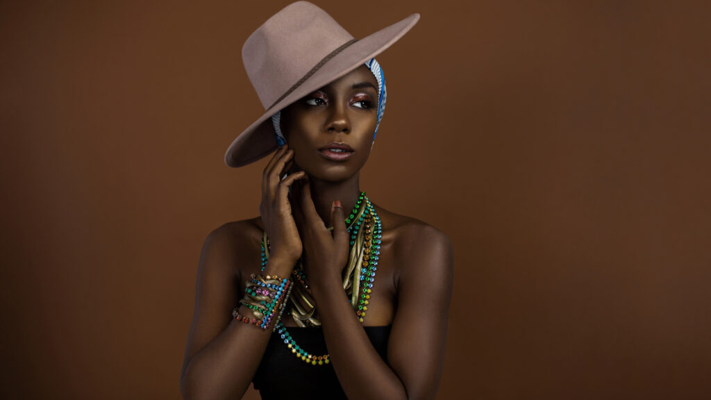 Serene looking young black female with beautiful makeup posing by herself in a studio wearing a black strapless top, blue & white head scarf, brown hat and gorgeous colorful jewelry.