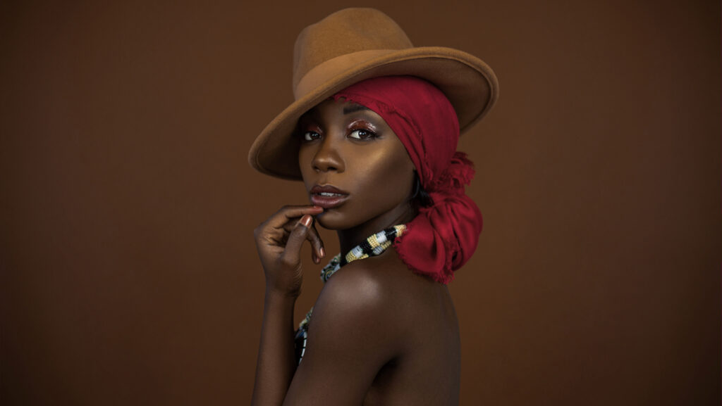 Sensual looking young black female with beautiful makeup posing by herself in a studio wearing a brown strapless top, red head scarf, brown hat and gorgeous colorful necklace.