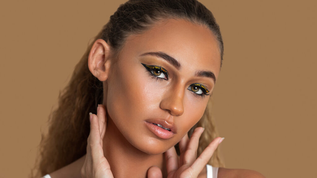 Modern beauty and self-expression. Lyla with long blond hair, light makeup, pastel eye shadow, touching her head, studio with pecan background.