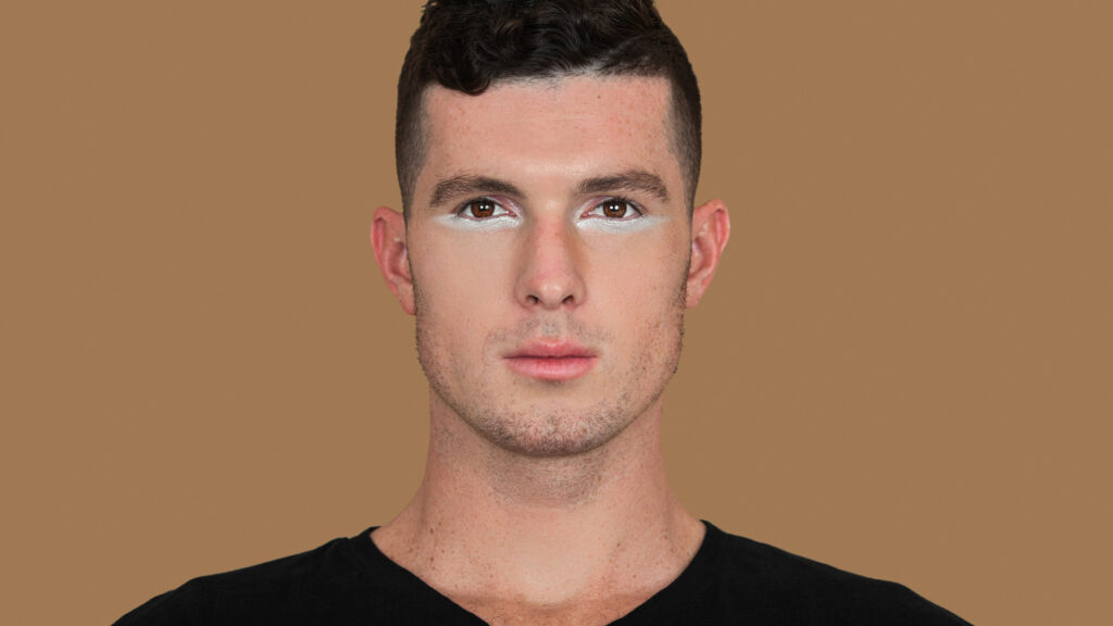 Modern beauty and self-expression. Ryan with short brown hair, white eyeshadow, light makeup, black shirt, studio with pecan background.