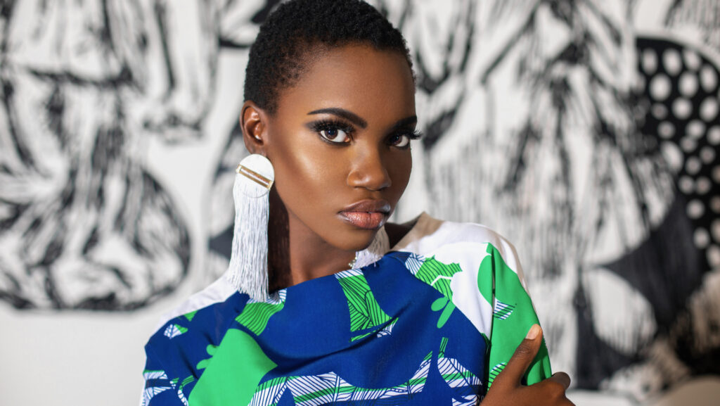 Pérez Art Museum Miami fashion editorial. An elegant young black woman with short black hair & beautiful makeup and dangling earrings posing by herself indoors is wearing a pretty dress.