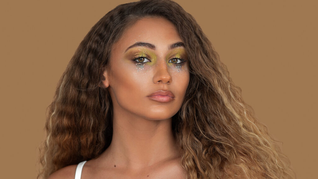 Modern beauty and self-expression. Lyla closeup with sparkling eye makeup, natural lipstick, long blond hair, studio with pecan background.