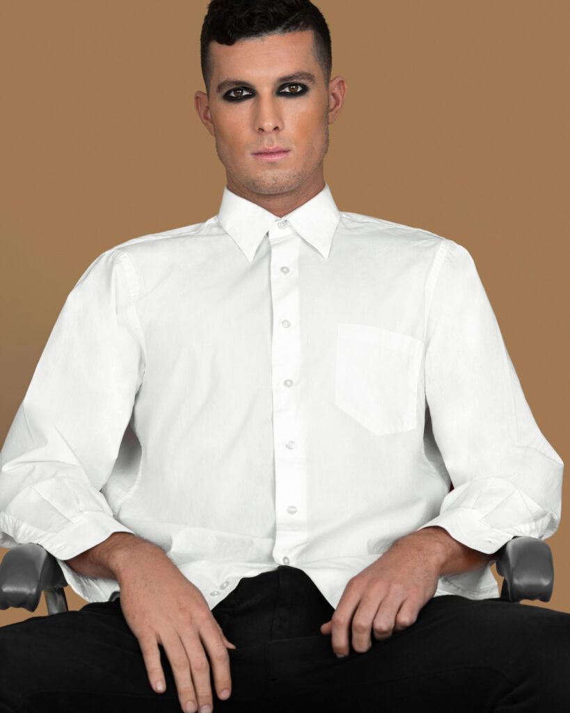 Modern beauty and self-expression. Ryan with short brown hair, dark eyeshadow, white shirt, black pants, studio with pecan background.