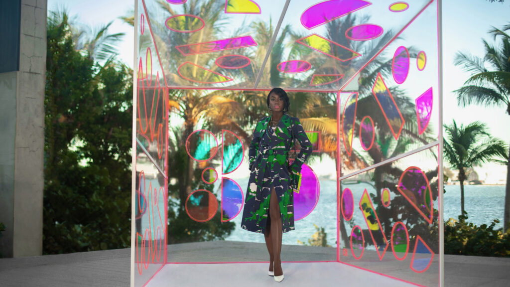 An elegant young black woman with long black hair & beautiful makeup & gorgeous jewelry posing by herself outdoors standing inside a glass box is wearing a colorful coat & hand bag white high heels.