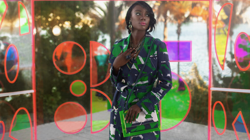 An elegant young black woman with long black hair & beautiful makeup & gorgeous jewelry posing by herself outdoors standing inside a glass box is wearing a colorful coat & hand bag.
