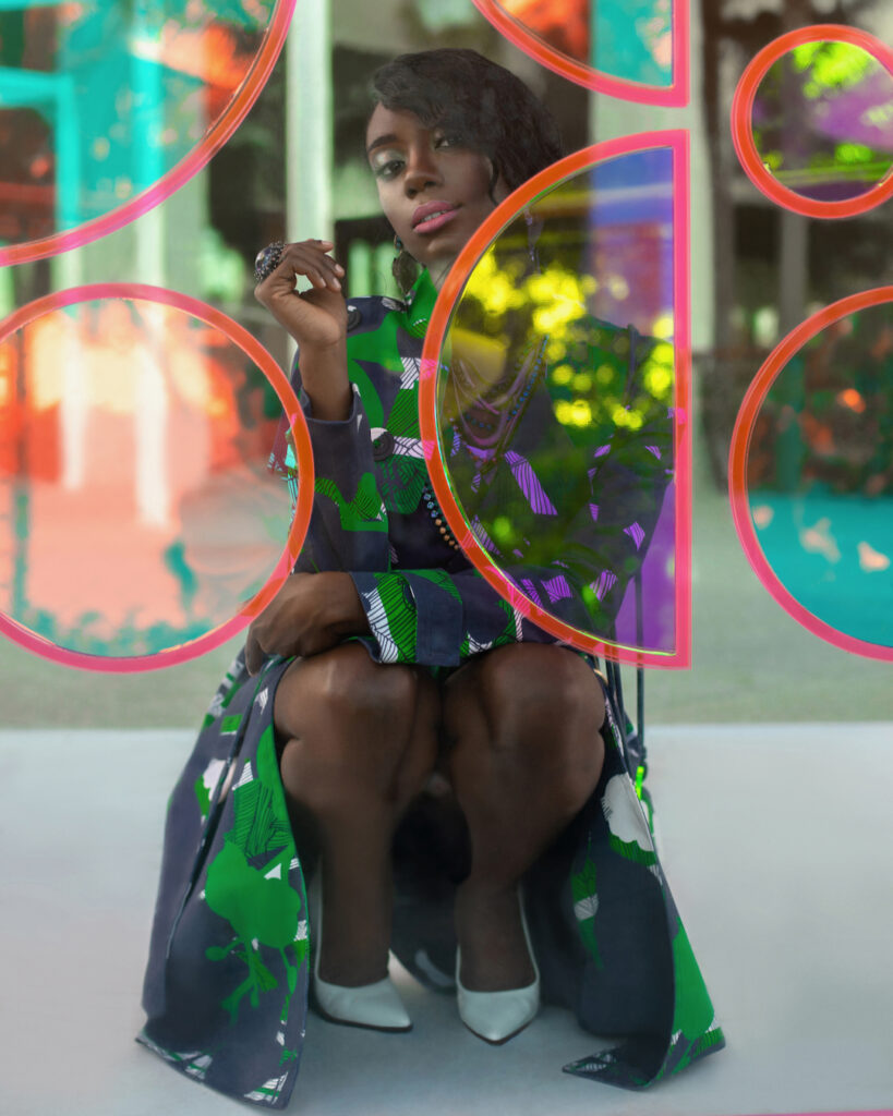 An elegant young black woman with long black hair & beautiful makeup & gorgeous jewelry posing by herself outdoors kneeling inside a glass box is wearing a colorful coat & hand bag white high heels.