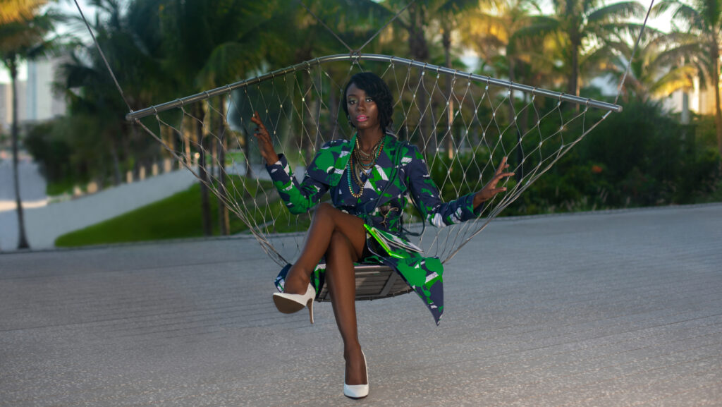 Pérez Art Museum Miami fashion editorial. An elegant young black woman with long black hair & beautiful makeup & gorgeous jewelry posing by herself outdoors sitting inside a swing is wearing a colorful coat & hand bag with white high heels.