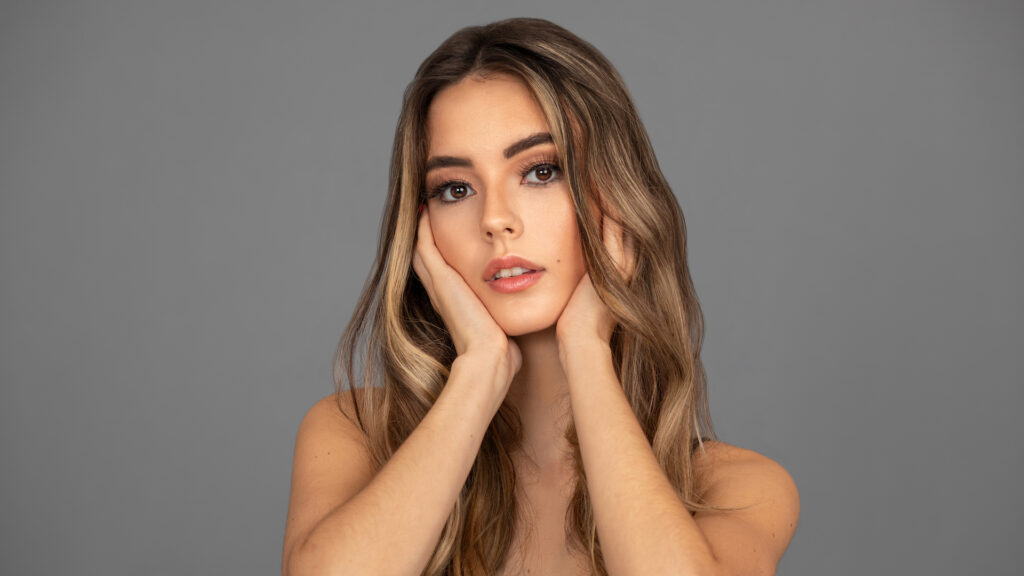 Closeup portrait of a sensual young Latina with long wavy hair, beautiful soft makeup and full eye brows posing by herself inside a studio with a gray background.