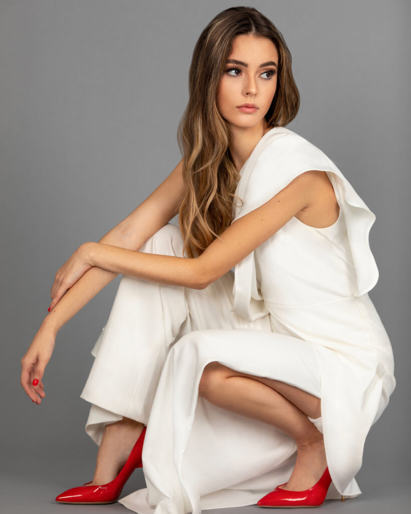Anti Demeure. Portrait of a beautiful young Latina with long wavy hair and soft makeup wearing a white evening dress with red high heels in a studio with gray background.