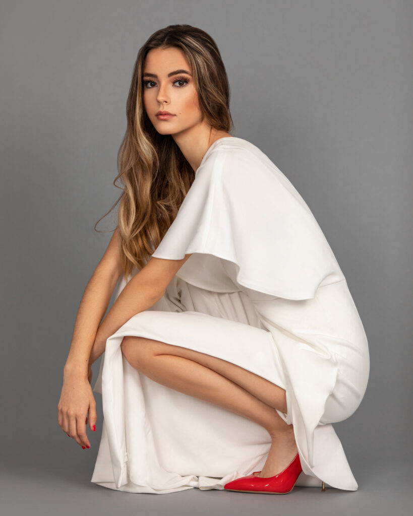 Portrait of a sensual young Latina with long wavy hair and soft makeup posing by herself inside a studio with a gray background wearing a white evening dress with red high heels.