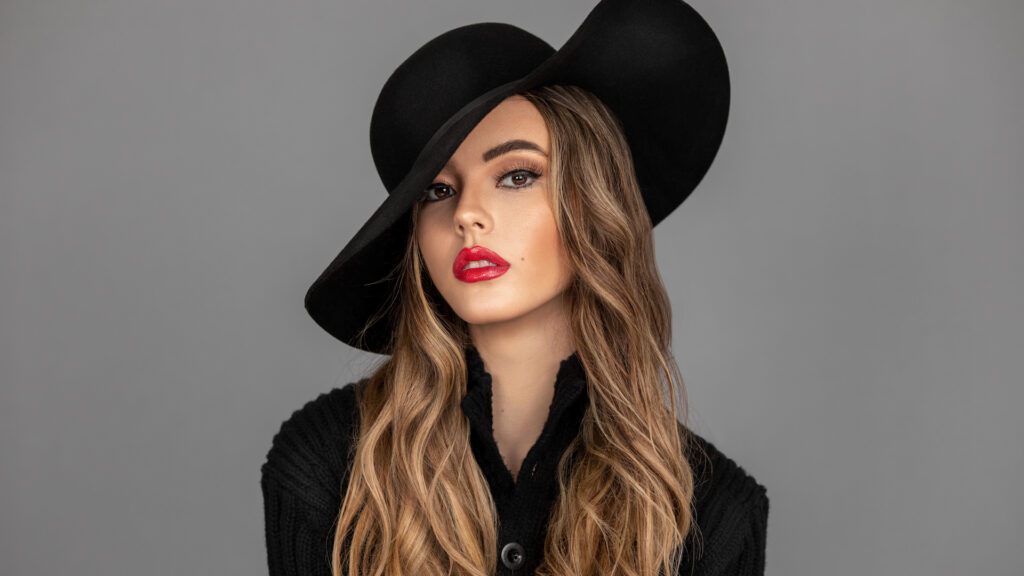 Anti Demeure. Closeup portrait of a sexy young Latina with long wavy hair, red lips, and full eyebrows wearing a black hat and sweater in a gray studio background.