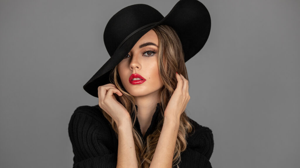 Closeup portrait of a sensual young Latina with long wavy hair, beautiful makeup with red lips and full eye brows posing by herself inside a studio with gray background wearing a black hat and sweater.