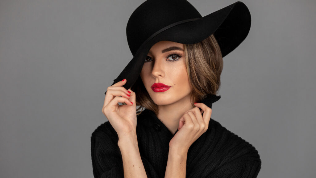 Closeup portrait of a shy young Latina with long wavy hair, beautiful makeup with red lips and full eye brows posing by herself inside a studio with gray background wearing a black hat and sweater.