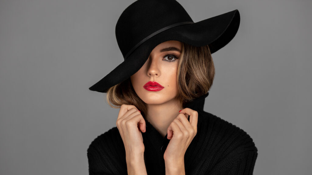 Closeup portrait of a young Latina with long wavy hair, beautiful makeup with red lips and full eye brows posing by herself inside a studio with gray background wearing a black hat and sweater.