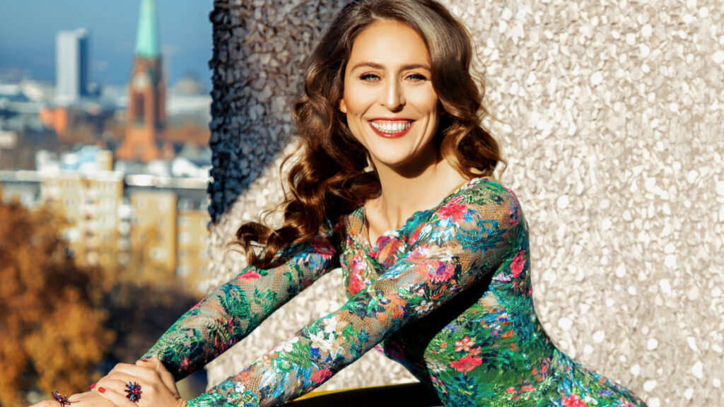 Portrait of a joyful white woman with long natural curly brown hair, beautiful makeup posing by herself outdoors on a terrace with a stone background wearing a colorful dress and jewelry.