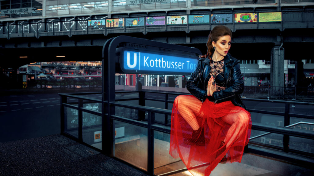 Portrait of a confident white woman with long natural curly brown hair, beautiful makeup posing by herself outdoors at Kottbusser Tor in Berlin wearing a red dress with a black leather jacket.