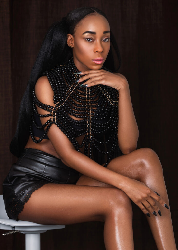 Portrait of a sensual young black woman with long hair and beautiful makeup sitting by herself on a bar chair inside a studio with a curtain background wearing black leather shorts with a sexy top.