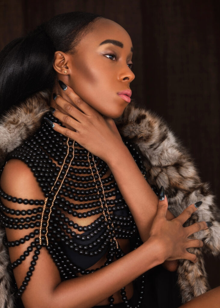 Portrait of a serene young black woman with long hair and beautiful makeup sitting by herself on a bar chair inside a studio with a curtain background wearing black pearls outfit and faux fur coat.