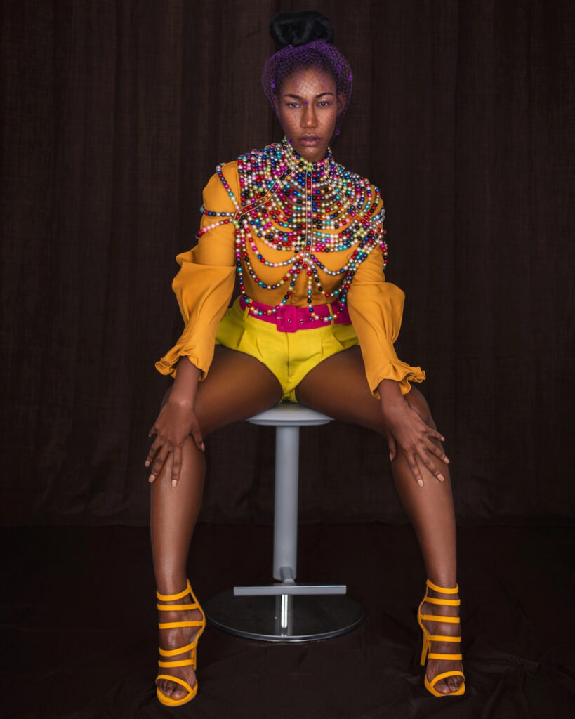 Portrait of a strong young black woman with long hair and beautiful makeup sitting by herself on a bar chair wearing yellow shorts, pink belt, orange blouse with colorful pearls and a purple veil.