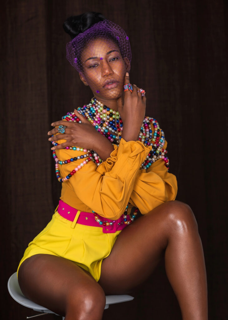 Closeup portrait of a sensual young black woman with long hair, beautiful makeup sitting by herself on a bar chair wearing yellow shorts, pink belt, orange blouse with colorful pearls and a veil.
