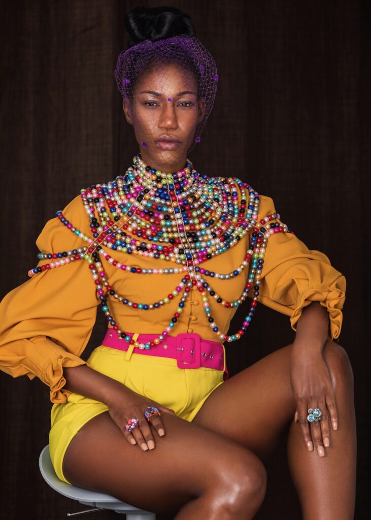 Closeup portrait of a confident young black woman with long hair, beautiful makeup sitting by herself on a bar chair wearing yellow shorts, pink belt, orange blouse with colorful pearls and a veil.