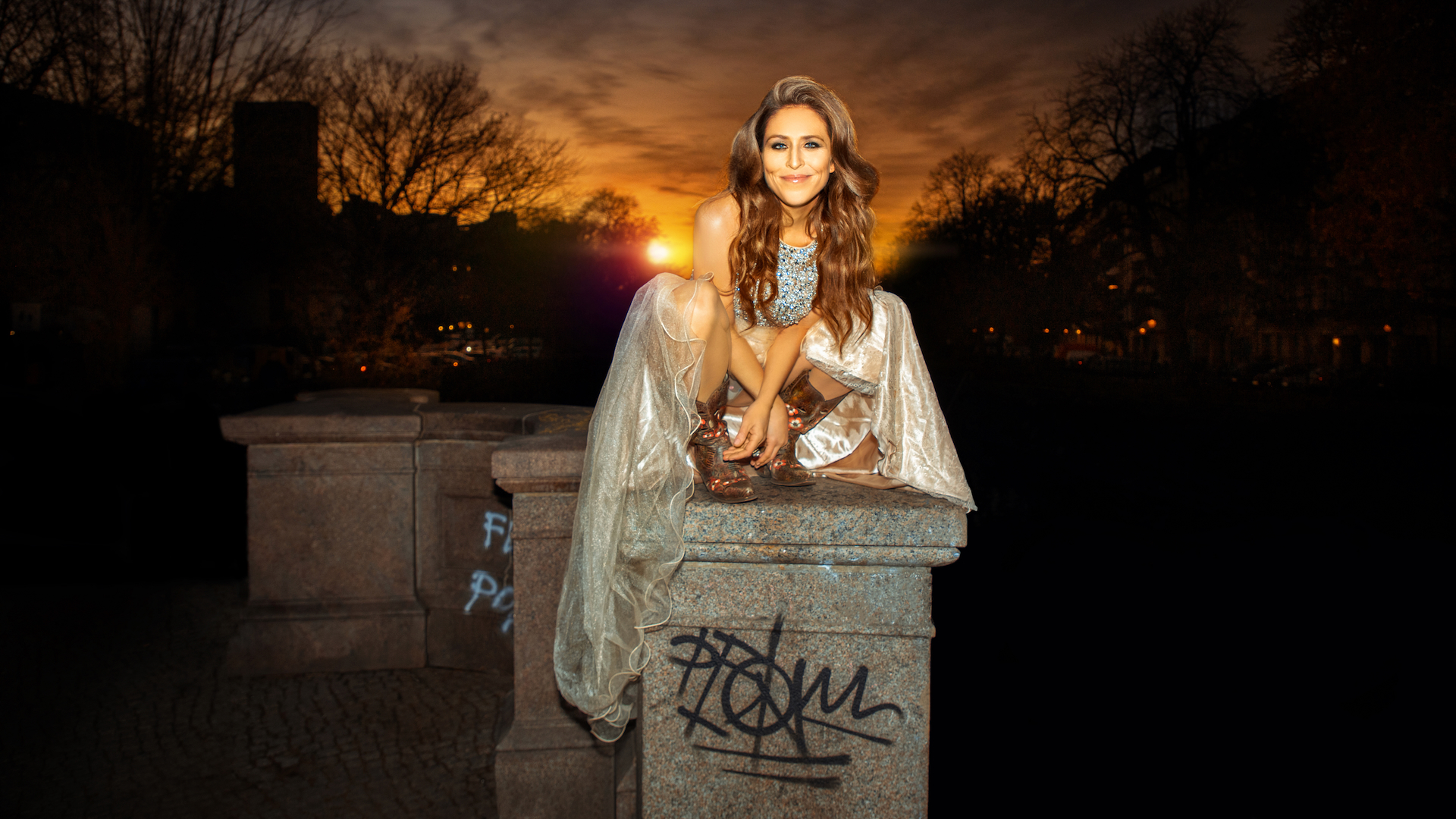 Britta Steffenhagen fashion editorial. Portrait of a beautiful white woman with long natural curly brown hair, beautiful makeup posing by herself outdoors sitting on top of a bridge in Berlin wearing a summer dress with leather boots.