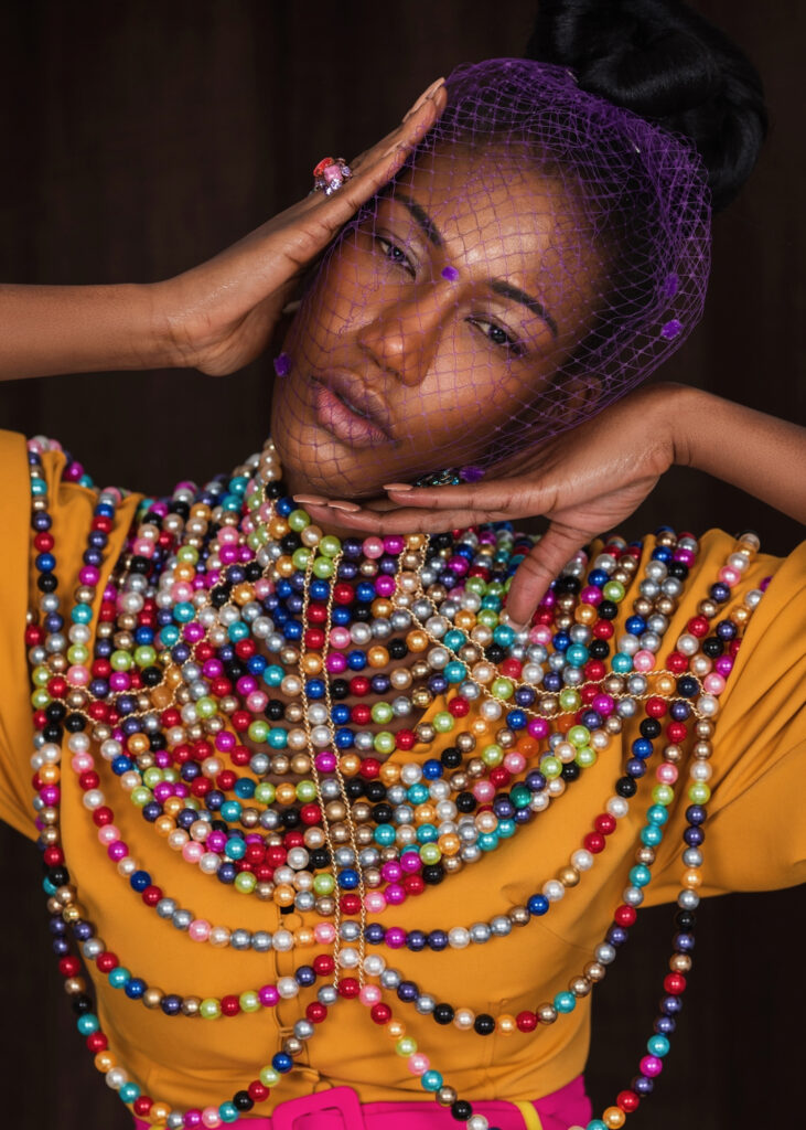 Closeup portrait of a young black woman with long hair, beautiful makeup posing by herself inside a studio with brown curtain background wearing an orange blouse with colorful pearls and purple veil.