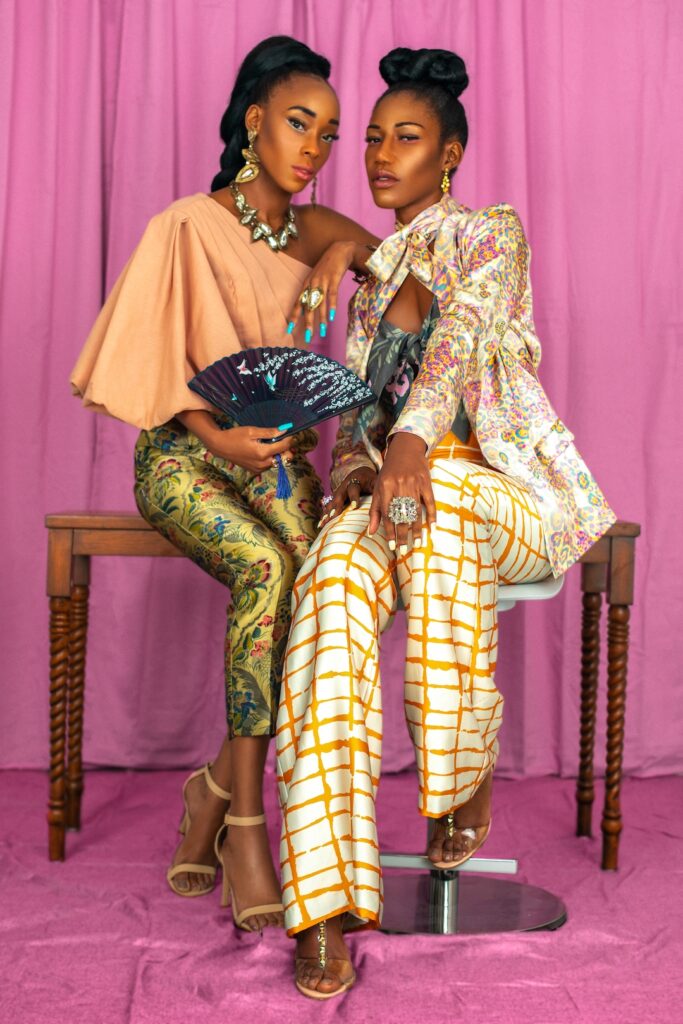 Portrait of two elegant black women with long hair, beautiful makeup posing together inside a studio with pink curtain background wearing elegant outfits with colorful jewelry and high heels.