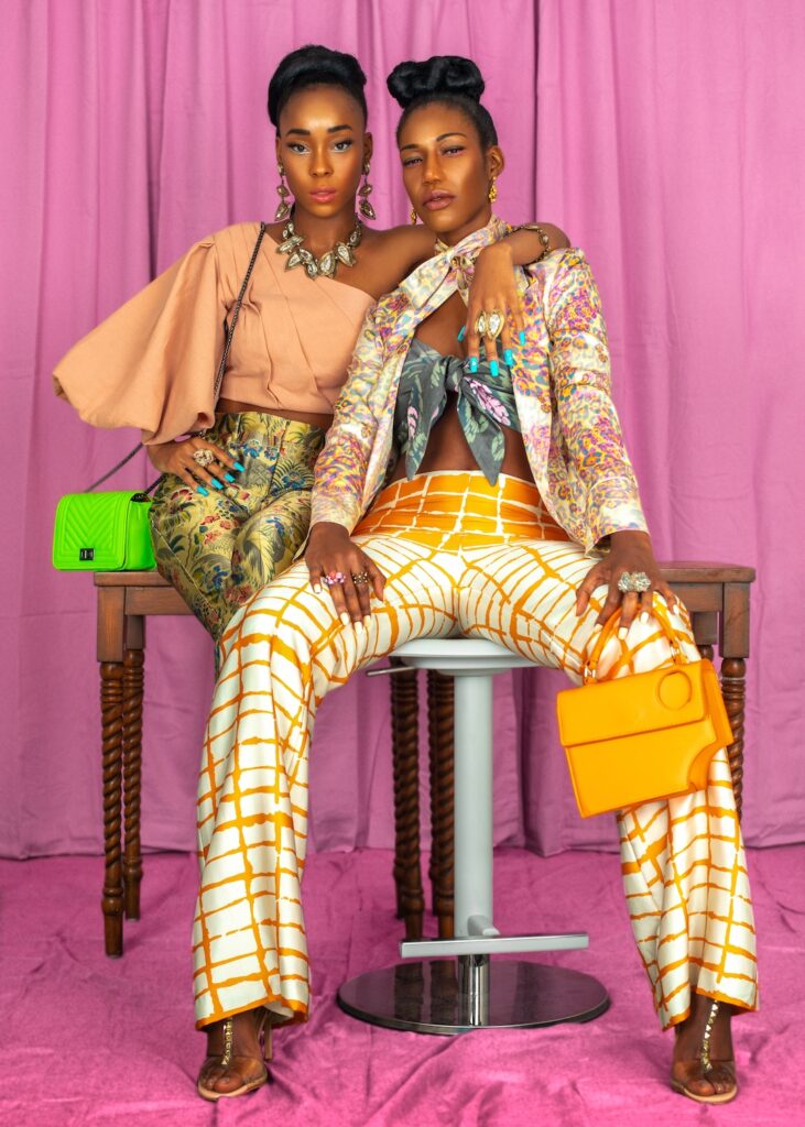 Portrait of two sexy black women with long hair, beautiful makeup posing together inside a studio with pink curtain background wearing elegant outfits with colorful jewelry and high heels.