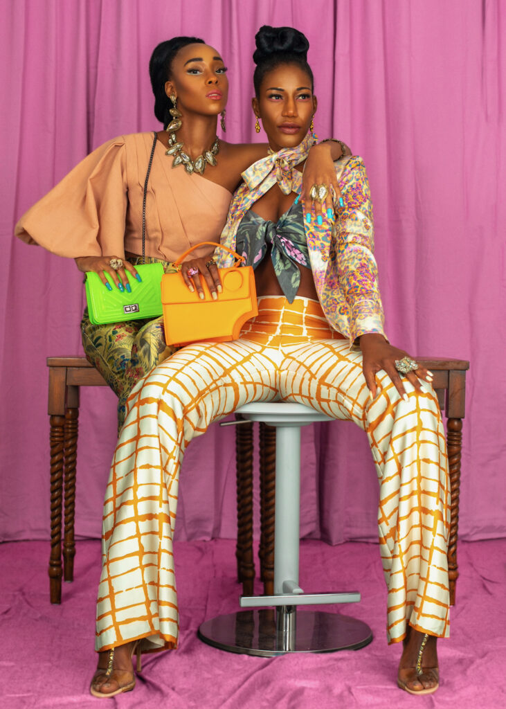 Portrait of two gorgeous black women with long hair, beautiful makeup posing together inside a studio with pink curtain background wearing elegant outfits with colorful jewelry and high heels.