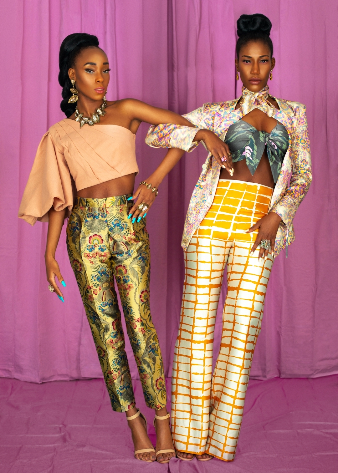 Trendy fashion for women. Portrait of two lovely black women with long hair, beautiful makeup posing together inside a studio with pink curtain background wearing elegant outfits with colorful jewelry and high heels.