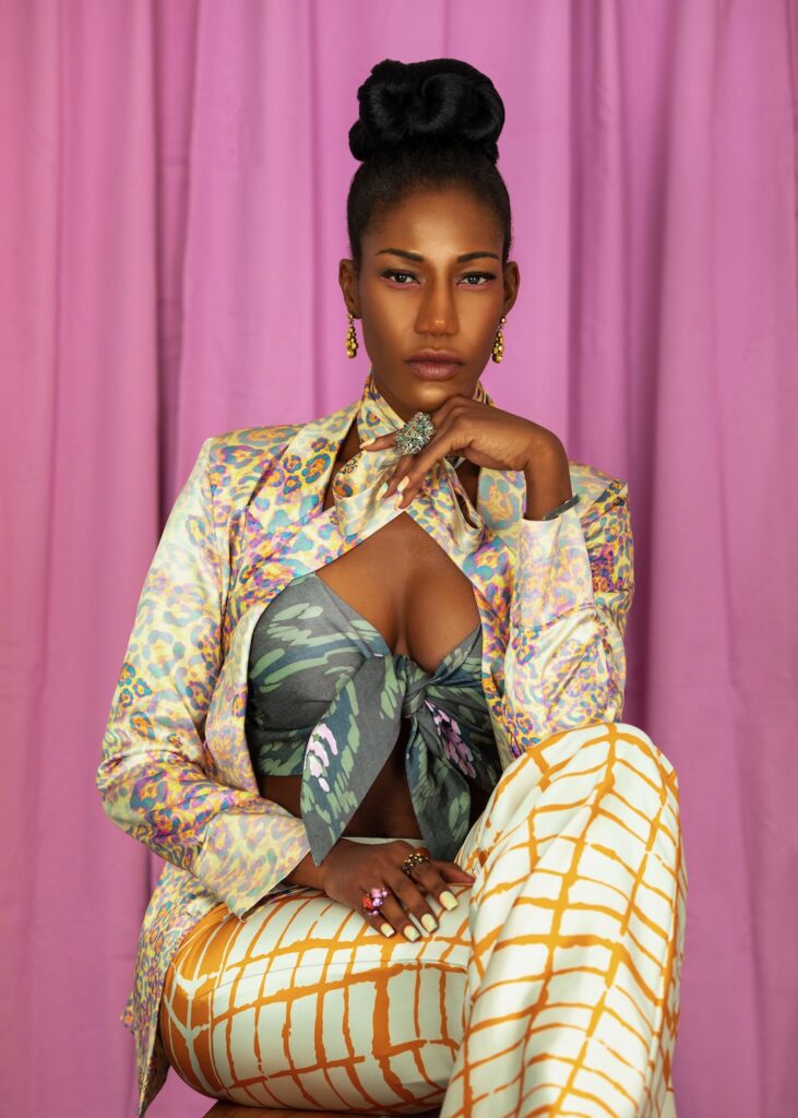 Closeup portrait of a pretty black woman with long hair, beautiful makeup posing by herself inside a studio with pink curtain background wearing an elegant satin outfit with colorful jewelry.