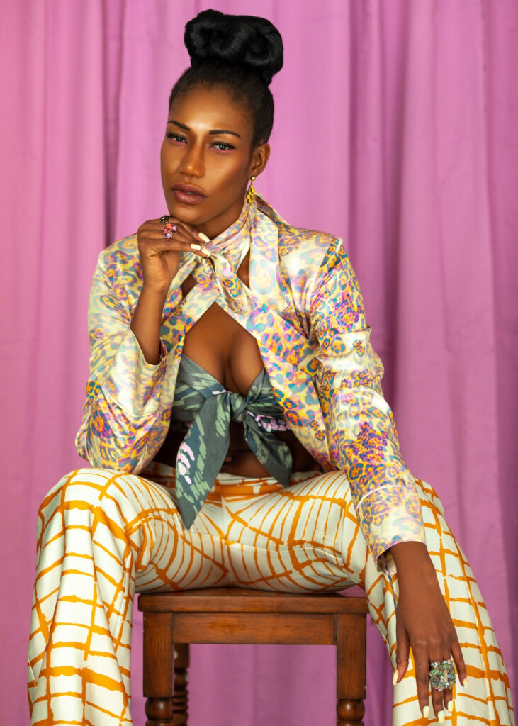 Portrait of an attractive black woman with long hair, beautiful makeup sitting by herself inside a studio with pink curtain background wearing an satin elegant outfit with colorful jewelry.