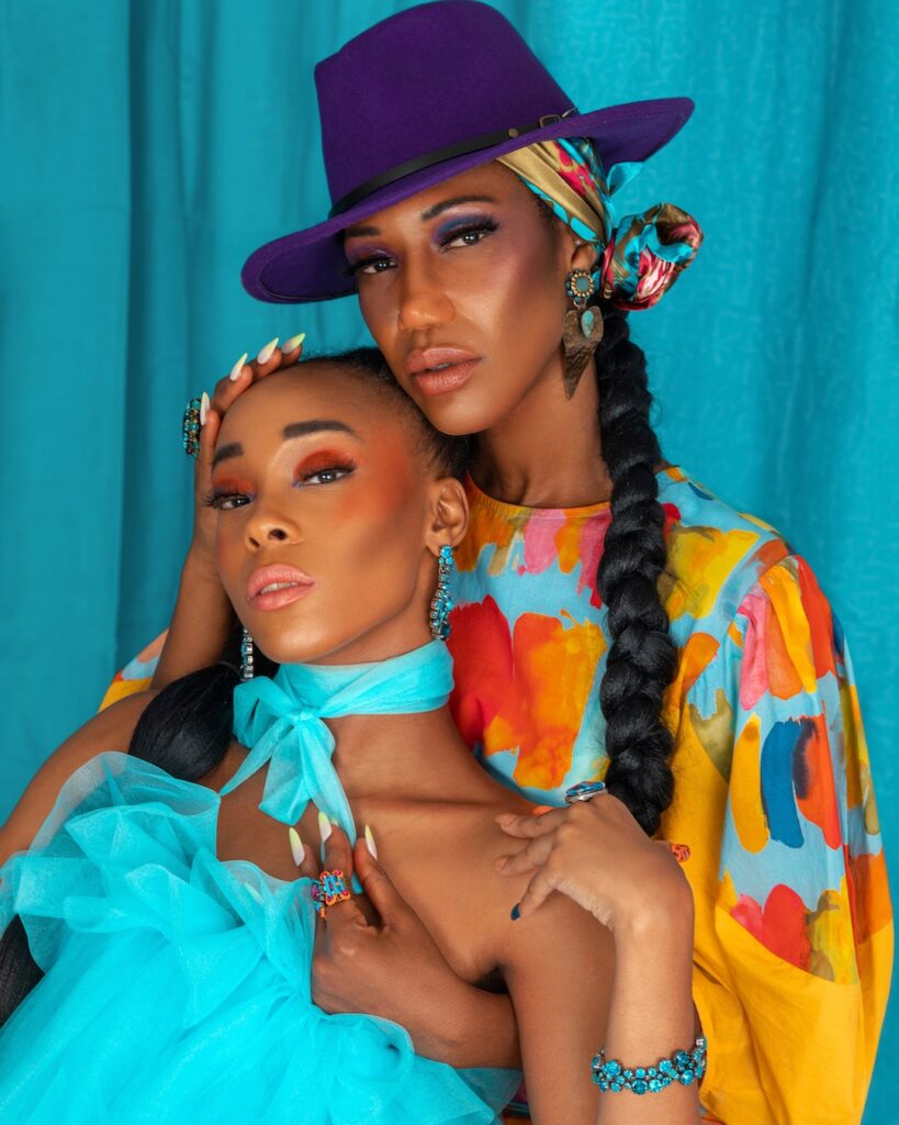 Closeup portrait of two beautiful black women with long hair, beautiful makeup posing together inside a studio with turquoise curtain background wearing elegant short dresses and jewelry.
