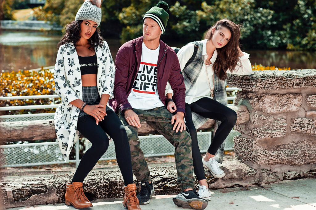 Urban fashion editorial photo of diverse young adults sitting on a wooden railing by a lake wearing stylish urban outfits.