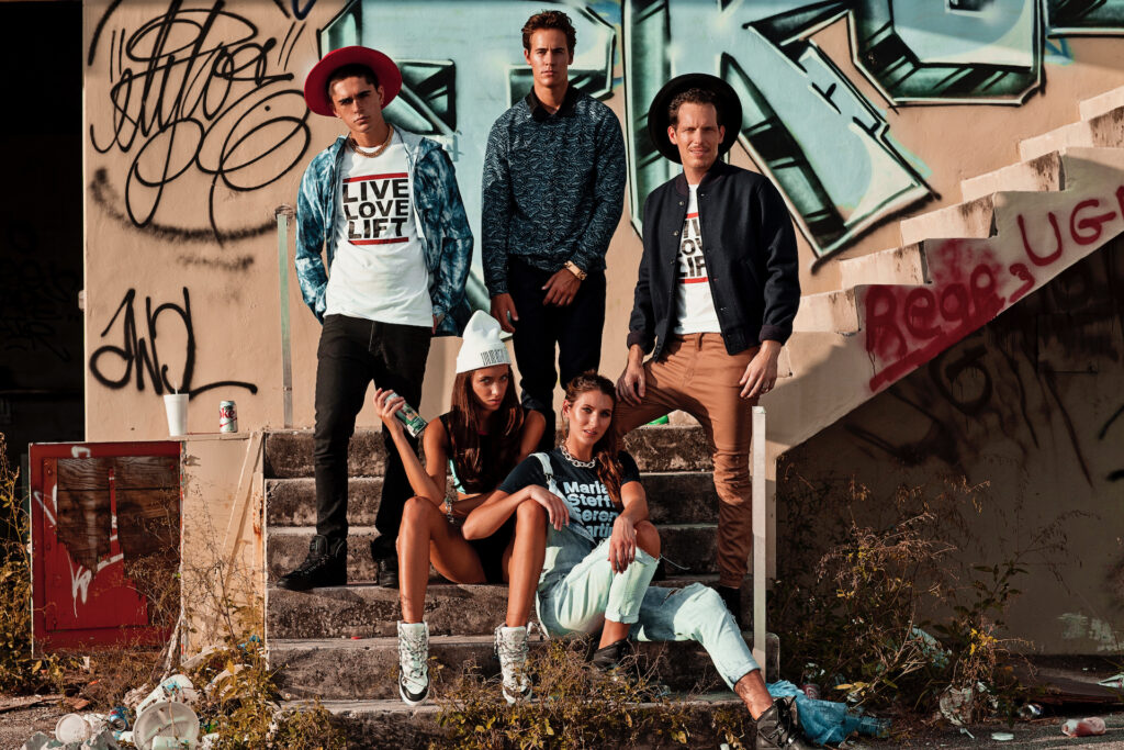 Group of five young adults wearing urban fashion outfits posing outdoors in front of an abandoned graffitied building in Miami.